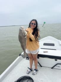 A smiling angler proudly displays a large Black Drum while fishing from a boat in a coastal waterway.