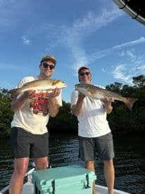 Two anglers proudly displaying their catches of Redfish while fishing on a boat in a scenic waterway.
