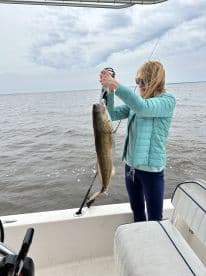 A woman proudly displaying a large fish while fishing on a boat, showcasing a successful catch in a scenic waterway.