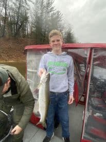A young angler proudly displaying a large trout while fishing from a boat in a scenic lake environment.