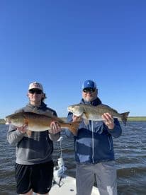 Two anglers proudly displaying their catches of Redfish while on a fishing charter, showcasing a sunny day on the water.
