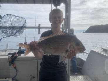 A young angler proudly displaying a Snapper caught while fishing on a boat, with a scenic ocean backdrop.