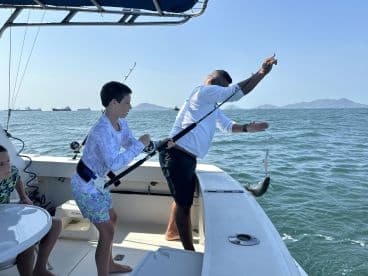 A young angler is actively reeling in a fish while another child observes on a fishing charter in open waters.