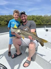 A young boy and his father proudly display a large Snook caught during their fishing trip on a sunny day.
