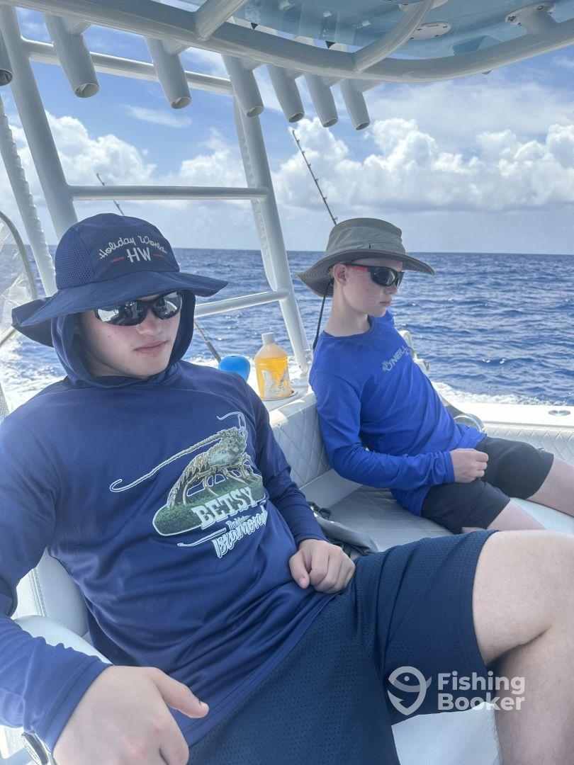 Two young anglers relaxing inside a boat, wearing sun protection gear, with a clear view of the ocean in the background.