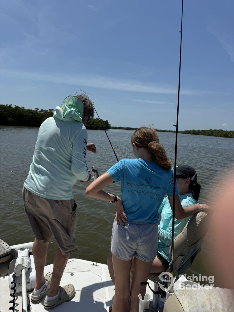 A group of young anglers engaged in fishing on a boat in a calm waterway, with one girl actively reeling in a catch.