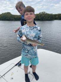 A young angler proudly displaying a Speckled Trout while fishing on a boat in a scenic estuary.