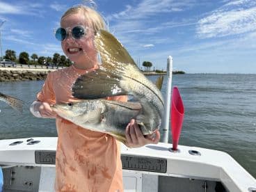 A young angler joyfully displaying a large Snook while fishing from a boat in a sunny coastal environment.