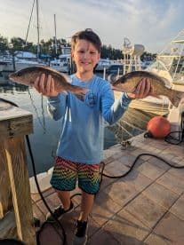 A young angler proudly displaying two Snapper catches at the dock, showcasing a successful fishing trip.