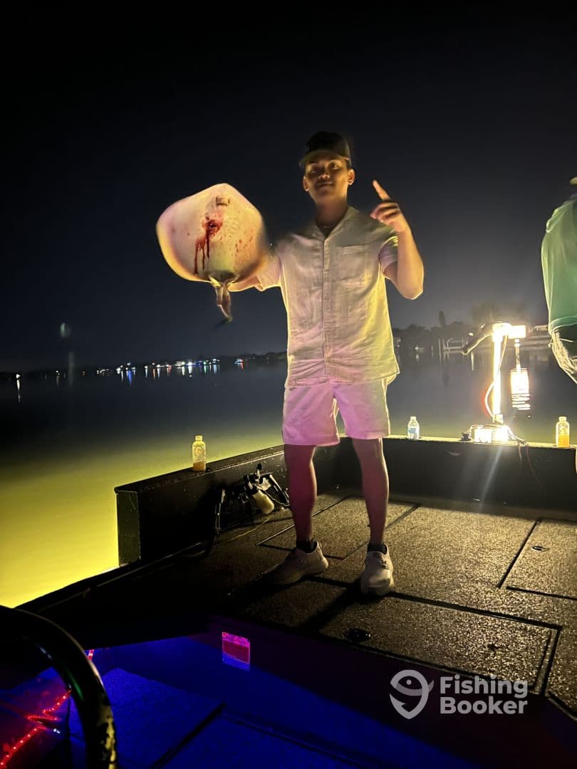 A young angler proudly displaying a caught fish on a boat during a night fishing trip, with a scenic waterfront backdrop.
