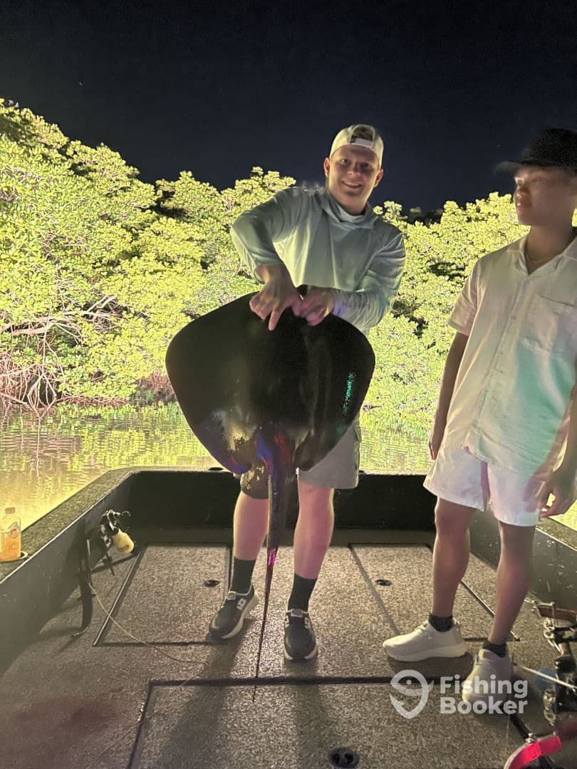 Two anglers on a boat at night, showcasing a large stingray caught during a fishing trip in a mangrove area.
