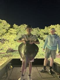 Young angler proudly displaying a large stingray aboard a boat at night, surrounded by lush mangroves.