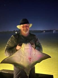 An angler proudly displaying a large stingray caught during a night fishing trip, illuminated by the water's glow.