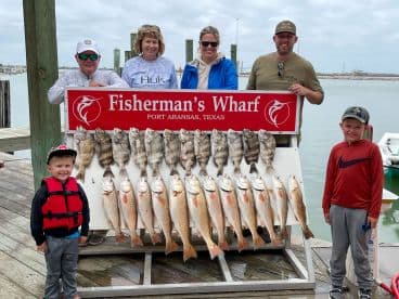 A family poses proudly in front of a catch board displaying an impressive array of fish, including Redfish and Black Drum, at Fisherman's Wharf in Port Aransas, Texas.