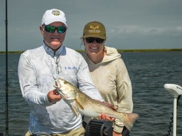 Two anglers proudly displaying a large Redfish while fishing in a scenic coastal environment.