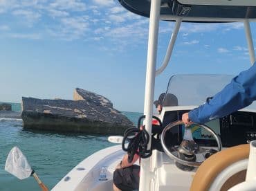 View from the helm of a boat navigating near a rocky outcrop in clear waters, showcasing the boat's interior and fishing electronics.