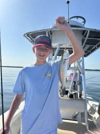A young angler proudly displaying a Speckled Trout while fishing on a boat in calm waters.