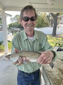 An older angler proudly displaying a Speckled Trout caught at a scenic dockside location.