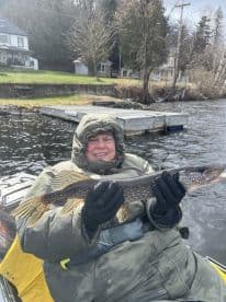 An angler proudly displaying a Northern Pike while fishing from a kayak on a serene lake, surrounded by lush greenery.