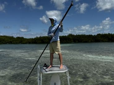 Angler standing on a polling platform, using a pole to navigate shallow waters in a mangrove environment.