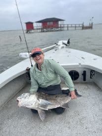 An angler proudly displaying a large Black Drum while seated on a fishing boat, with a scenic fishing lodge in the background.