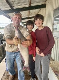 A proud angler holding a large catfish with two young boys at a fishing dock, showcasing a successful family fishing trip.