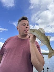 An angler playfully poses with a freshly caught Walleye, showcasing the beautiful lake backdrop on a sunny day.
