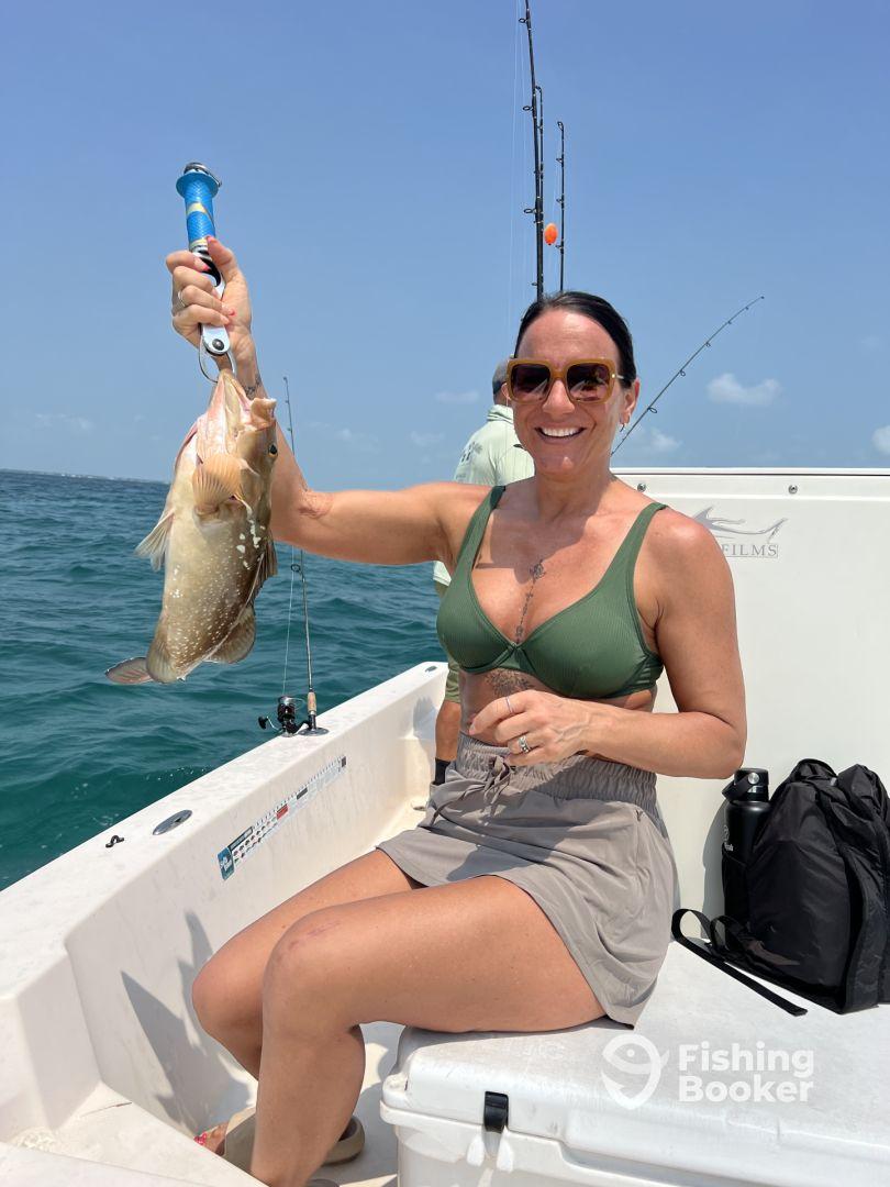 An angler proudly displaying a large Grouper while fishing on a boat in clear blue waters.