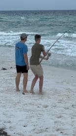 Two young anglers fishing on the beach, with one reeling in a catch while the waves crash in the background.
