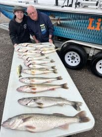 A young angler and an adult proudly display a variety of freshly caught fish, including several Walleye, arranged neatly on a table beside a fishing boat.