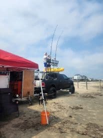 A fishing setup on the beach featuring a truck with a kayak on top and fishing rods, with a person adjusting equipment.