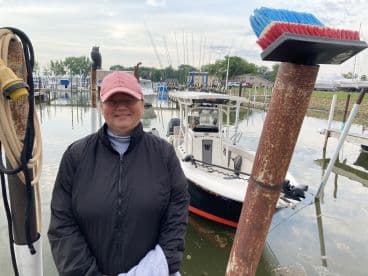 A smiling woman stands at the dock beside a fishing boat, ready for a day on the water. The marina features multiple boats and fishing gear in the background.