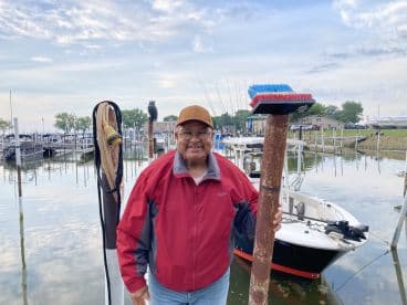 An older gentleman poses with a cleaning brush at the marina, showcasing a fishing boat in the background, surrounded by calm waters and a scenic harbor.