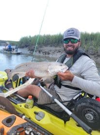 Angler proudly displaying a large striped bass while kayaking in a coastal marsh environment.