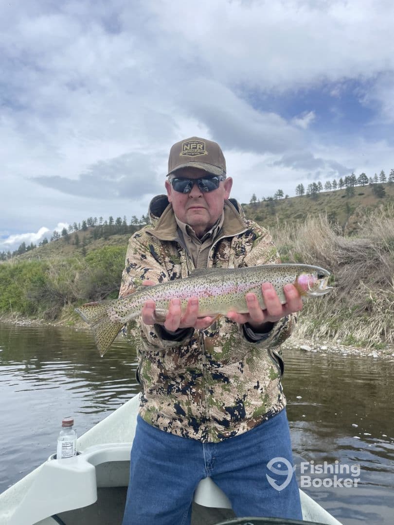 An angler proudly displaying a Rainbow Trout while fishing from a boat in a scenic river setting.