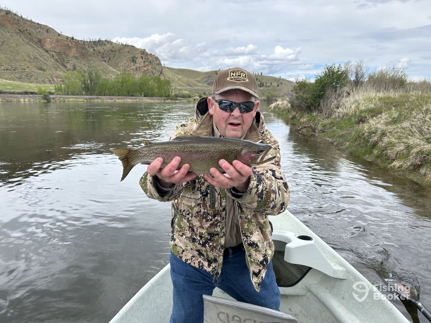 An angler proudly displaying a Rainbow Trout while fishing in a scenic river setting, surrounded by lush greenery and hills.