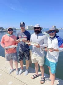 Four anglers proudly displaying their catch of Speckled Trout at the dock, enjoying a sunny day on the water.