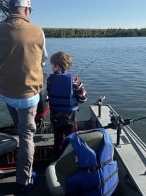 A young boy in a life jacket is fishing alongside an adult on a boat, enjoying a day on the water.