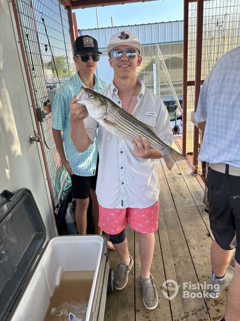 A young angler proudly displaying a striped bass while standing on the dock, with friends nearby, after a successful fishing trip.