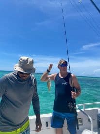 Two anglers on a boat in clear waters, with one proudly displaying a caught fish while the other prepares for the next catch.
