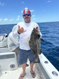 Angler proudly displaying a large Grouper while fishing on a boat in clear blue waters.