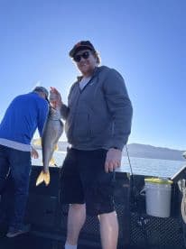 Angler proudly displaying a large fish on a boat during a sunny fishing trip, with scenic mountains in the background.