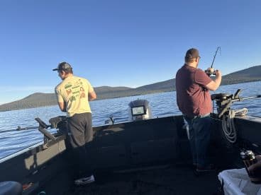 Two anglers actively fishing from a boat on a serene lake, surrounded by mountains under a clear blue sky.