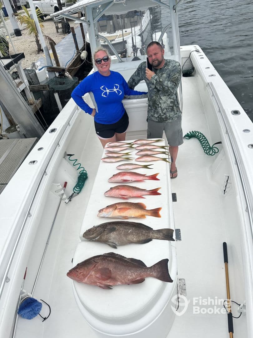 Two anglers proudly display a variety of freshly caught fish, including Snapper and Grouper, arranged on the boat's cleaning station after a successful fishing trip.