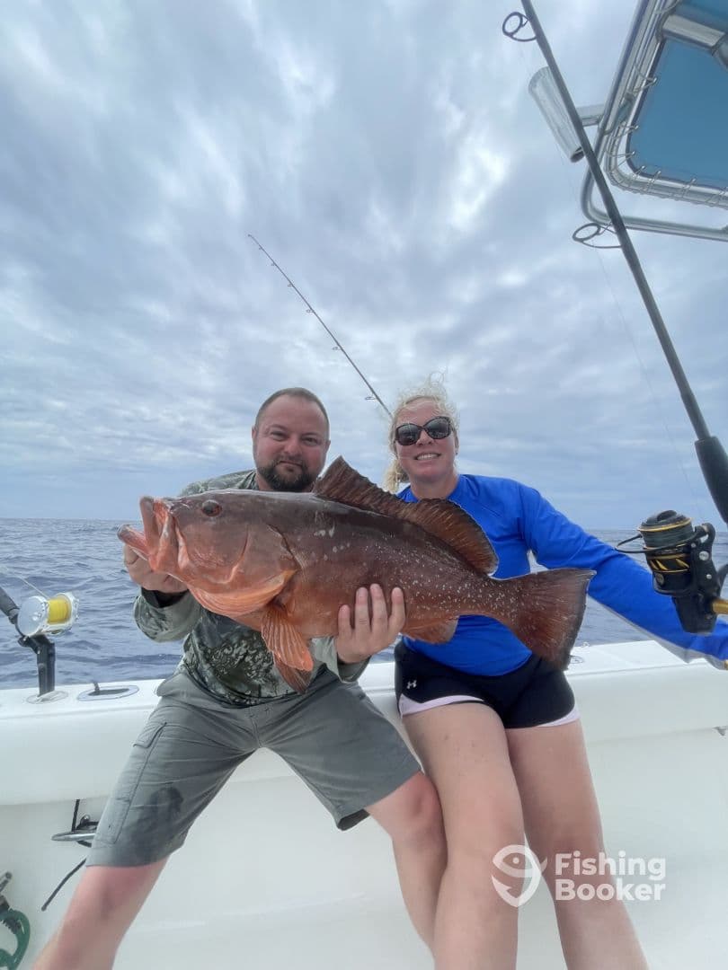 A family enjoying a successful fishing trip, proudly displaying a large Red Grouper on a boat in open water.