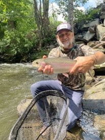 An angler proudly displaying a Rainbow Trout caught in a scenic river setting, surrounded by lush greenery.