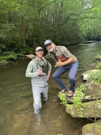 Two anglers enjoying a day of fishing in a scenic stream, surrounded by lush greenery.