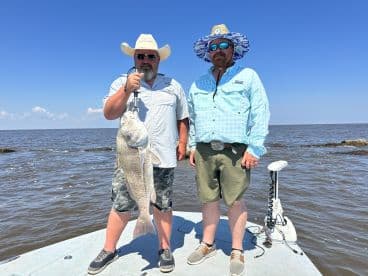 Two anglers proudly displaying a large Redfish on a fishing boat in a coastal environment.