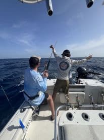 Two anglers actively fishing on a boat, with one reeling in a catch while the other assists, showcasing a dynamic offshore fishing experience.