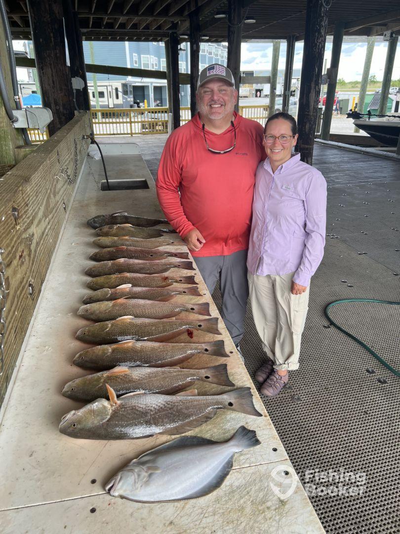 Two anglers proudly display their catch of various fish, including flounder and trout, arranged neatly on a cleaning table at a fishing dock.